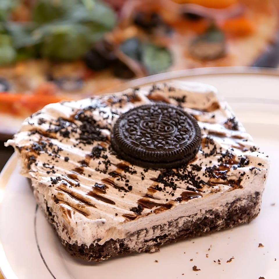 A slice of Oreo cheesecake with whipped cream, chocolate drizzle, and an Oreo cookie on top.