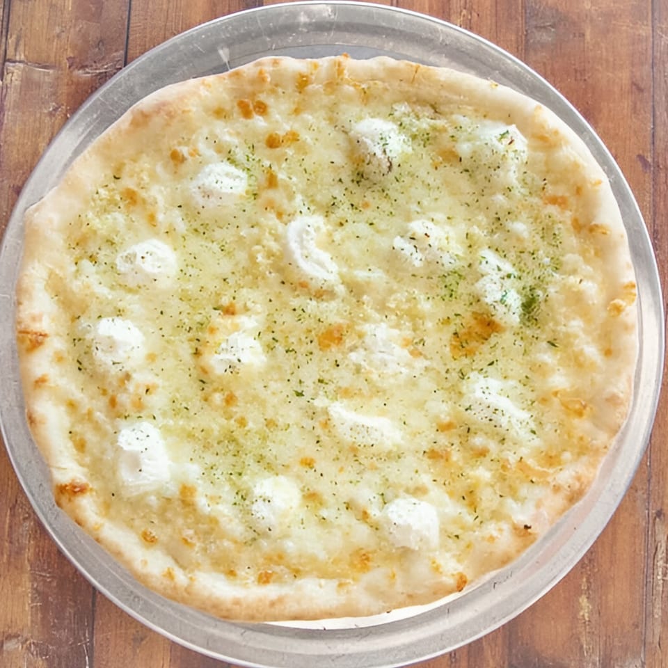 A white pizza with dollops of ricotta cheese and green herbs on a metal pizza pan.